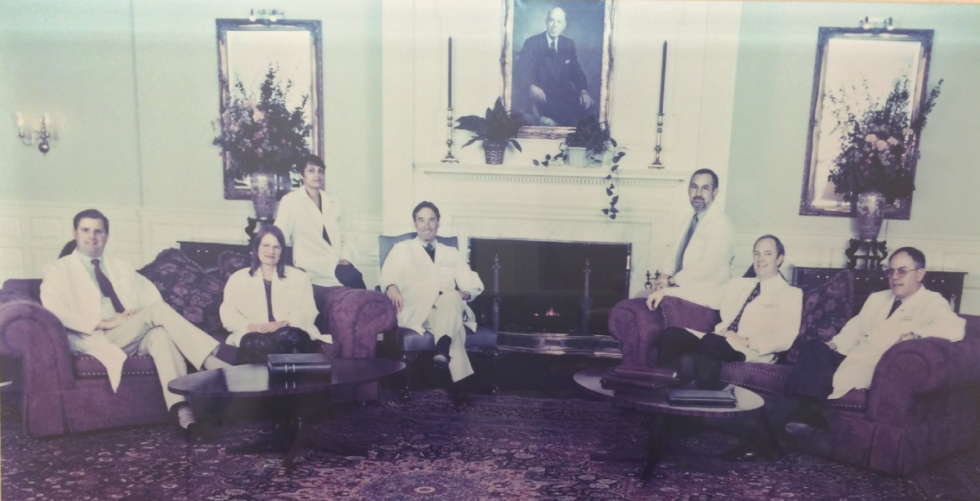 Six doctors in white coats sit on sofas in a formal room with a fireplace and large portrait.