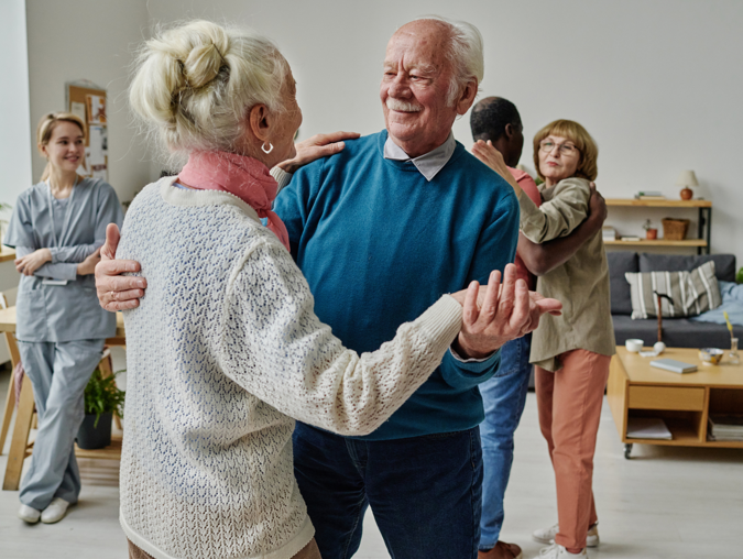 Elderly couples dancing together in a cozy room, caregiver watching and smiling.