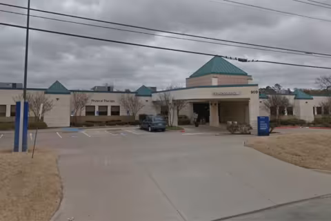 Single-story hospital entrance with blue accents, cloudy sky overhead, and parked vehicles.