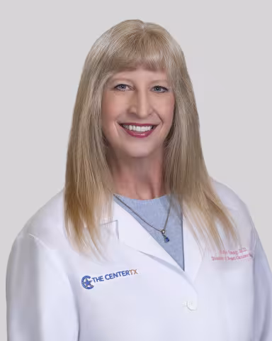Person in white lab coat with long blonde hair, blurred face, neutral background.