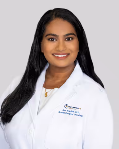 Smiling woman with long dark hair in a white medical coat, neutral background.