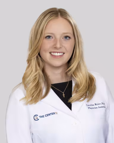 Woman with blonde hair smiling, wearing a white medical coat against a plain background.