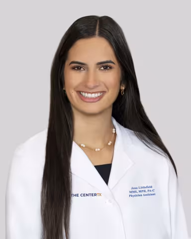 Woman smiling, wearing a white medical coat, with long dark hair, on a gray background.