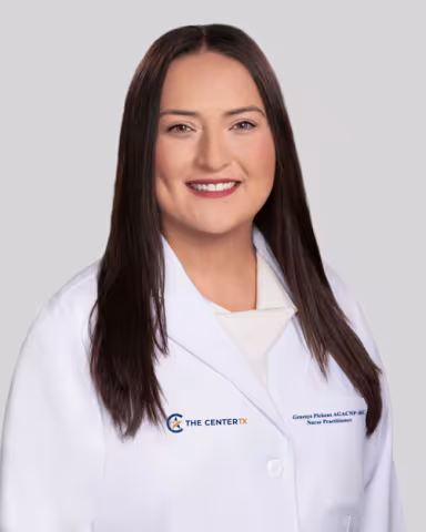 Woman smiling, wearing a white medical coat against a light gray background.