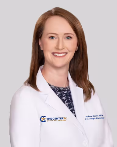 Smiling woman in a white medical coat with brown hair against a light gray background.