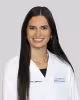 Woman smiling, wearing a white medical coat, with long dark hair, on a gray background.