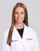 Woman with straight brown hair wearing a white medical coat, smiling against a gray background.