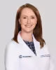 Smiling woman in a white medical coat with brown hair against a light gray background.
