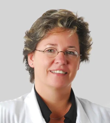 Person with short, wavy hair wearing a white coat and black shirt against a gray background.
