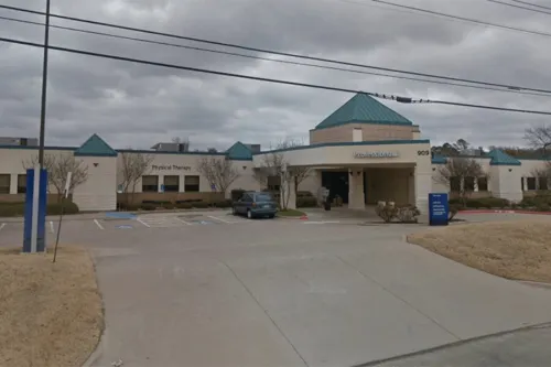Single-story hospital entrance with blue accents, cloudy sky overhead, and parked vehicles.