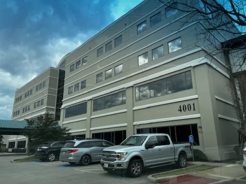 Four-story office building with parked cars in front under a cloudy sky.