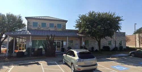 Stone building with large windows, trees in front, cars in a parking lot with handicap spaces.