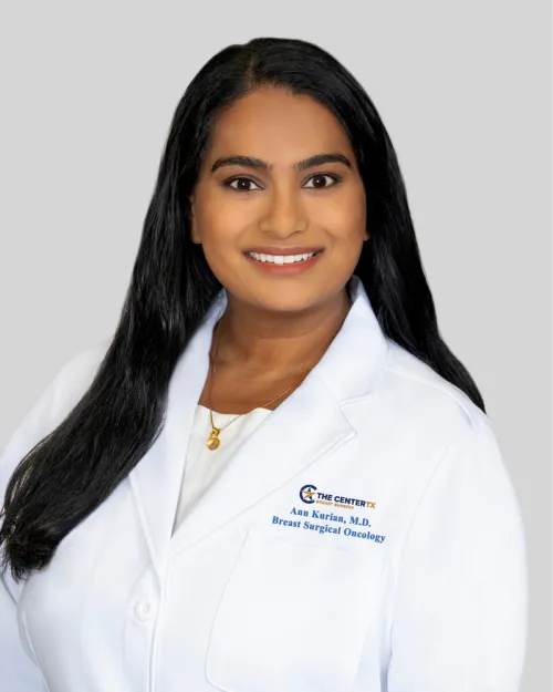 Smiling woman with long dark hair in a white medical coat, neutral background.