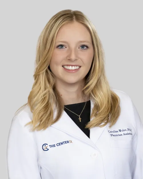 Woman with blonde hair smiling, wearing a white medical coat against a plain background.