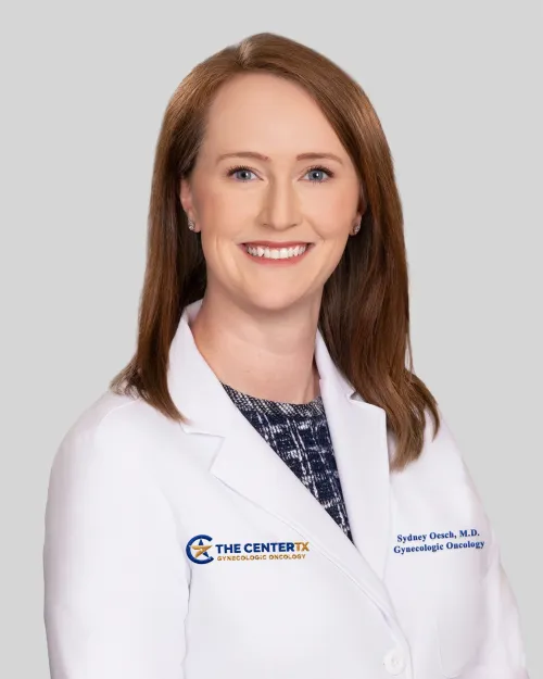 Smiling woman in a white medical coat with brown hair against a light gray background.